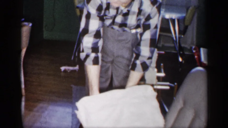In a 1959 Michigan home interior, a man in a checkered shirt stands by folding chairs while handling a white cloth.