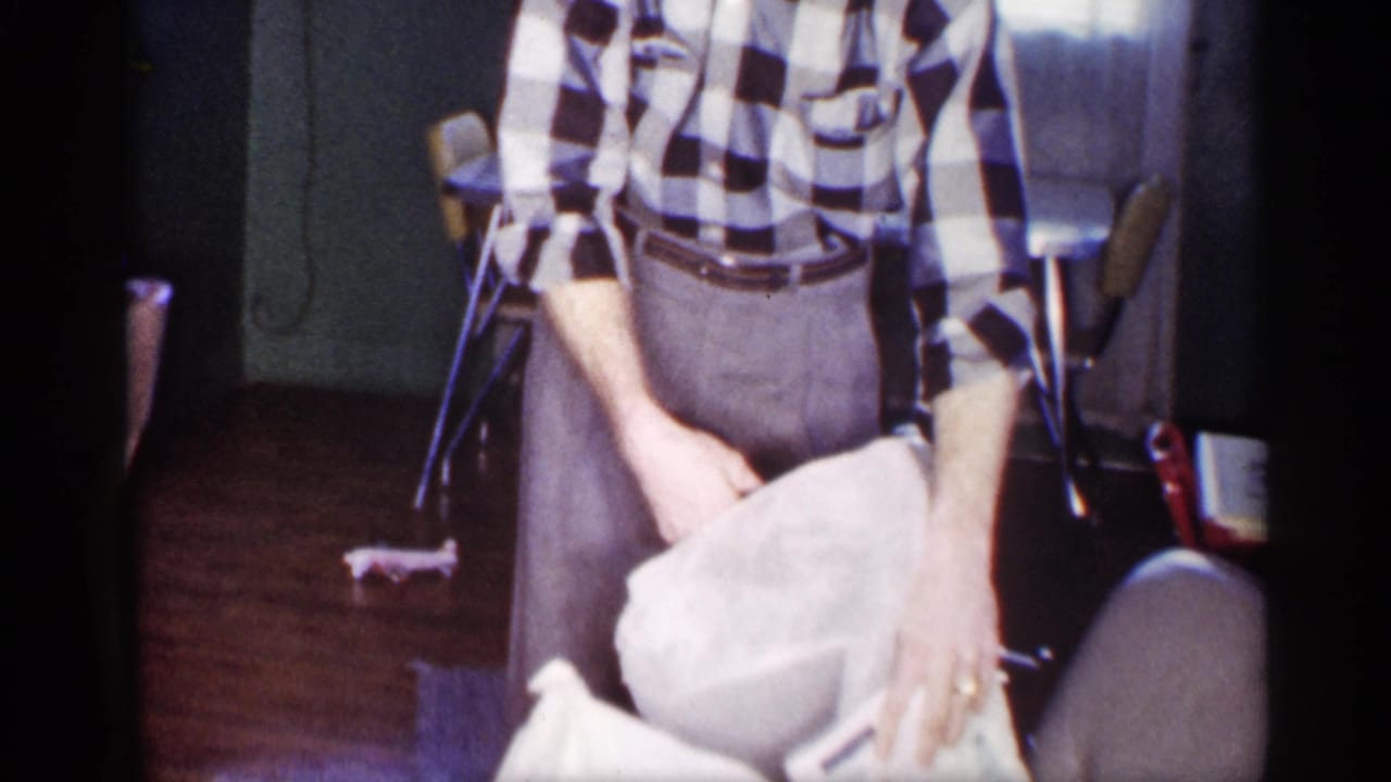 The man in a black-and-white checkered shirt holds a white cloth in a softly lit Michigan home interior.