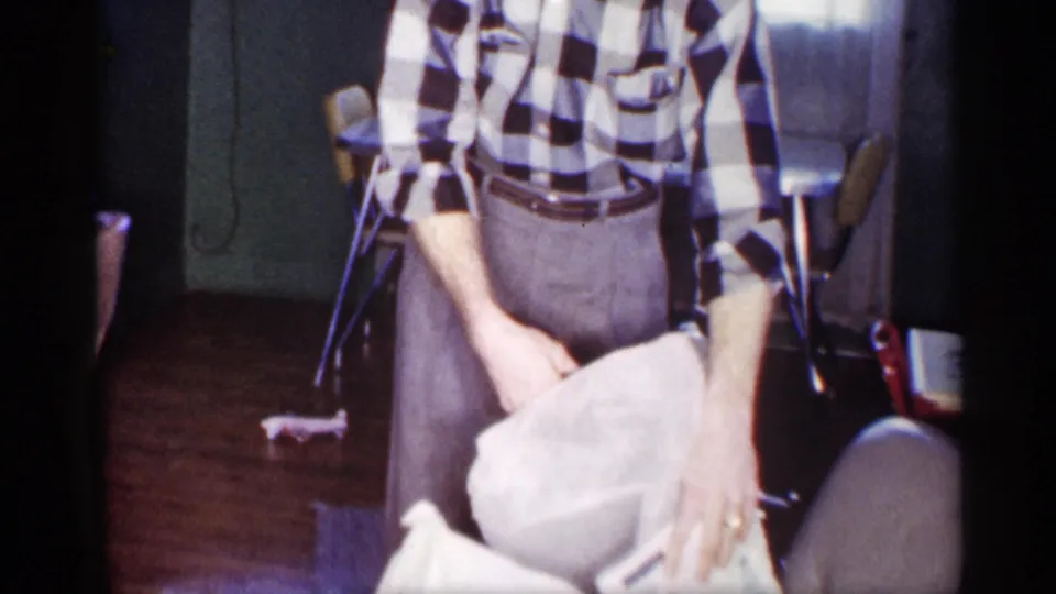 The man in a black-and-white checkered shirt holds a white cloth in a softly lit Michigan home interior.