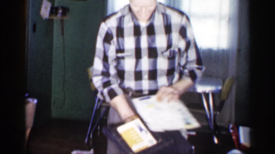 A man in a checkered shirt holds a white cloth and a yellow booklet or package in a 1959 Michigan home setting.