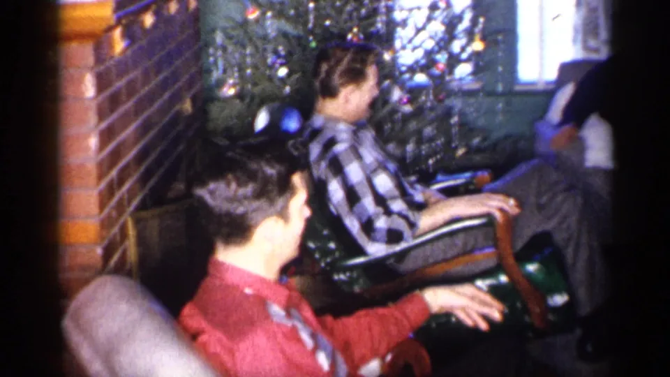 Two adult men sit in a 1959 Michigan home near a decorated Christmas tree, with one man turning and gesturing from his chair.