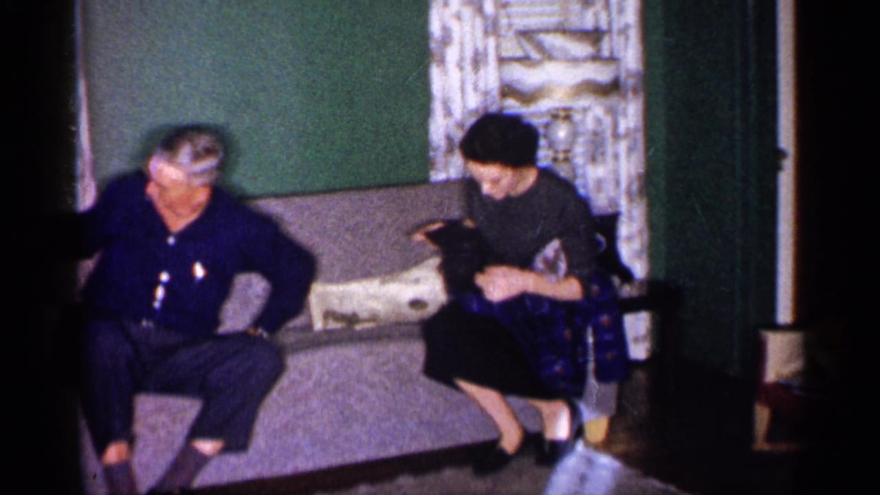 1959 Michigan home-movie frame showing an older man and woman seated on a couch in a modest living room.