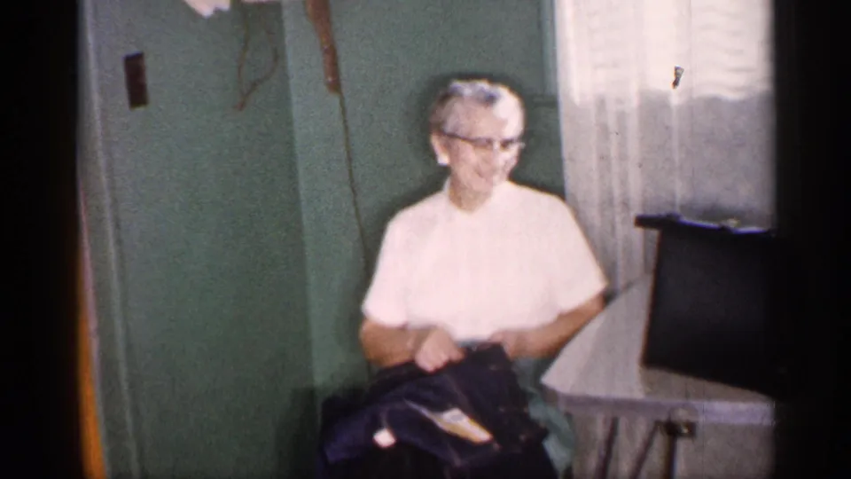 1959 Michigan home interior frame showing an older woman seated beside a small table near curtains and a cabinet.