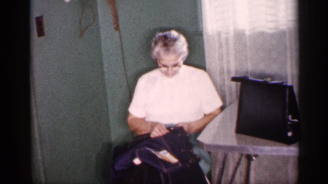 Older woman sits indoors looking down at a dark bag near a window with sheer curtains.