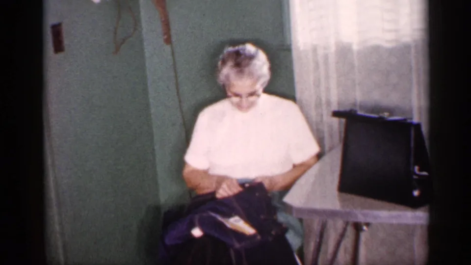 In a 1959 Michigan home interior, an older woman in a light blouse sits beside green walls and sheer curtains while looking into a dark bag.