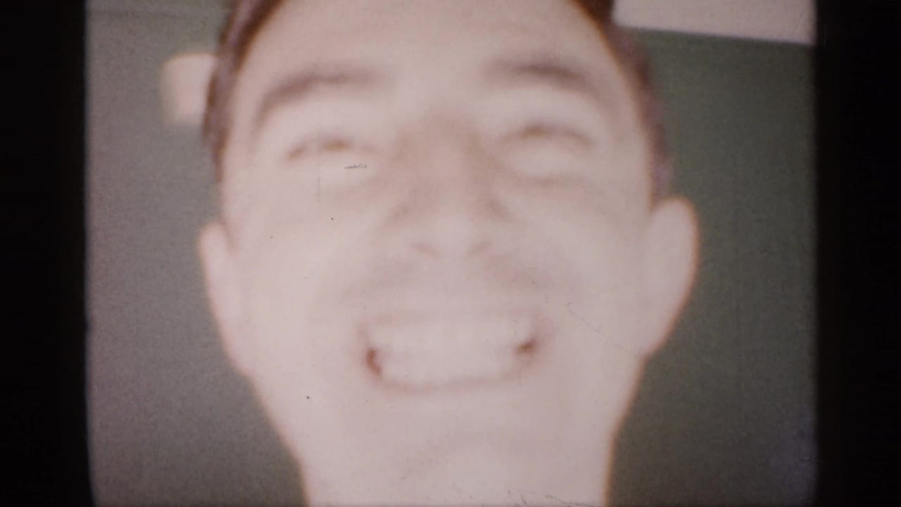 A young man smiles very close to the 8mm camera inside the Michigan home, with the frame heavily overexposed and softly focused.