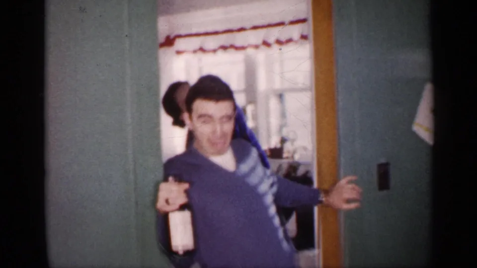 A young man in a blue V-neck sweater holds a bottle and gestures playfully from a doorway in the 1959 Michigan home interior.