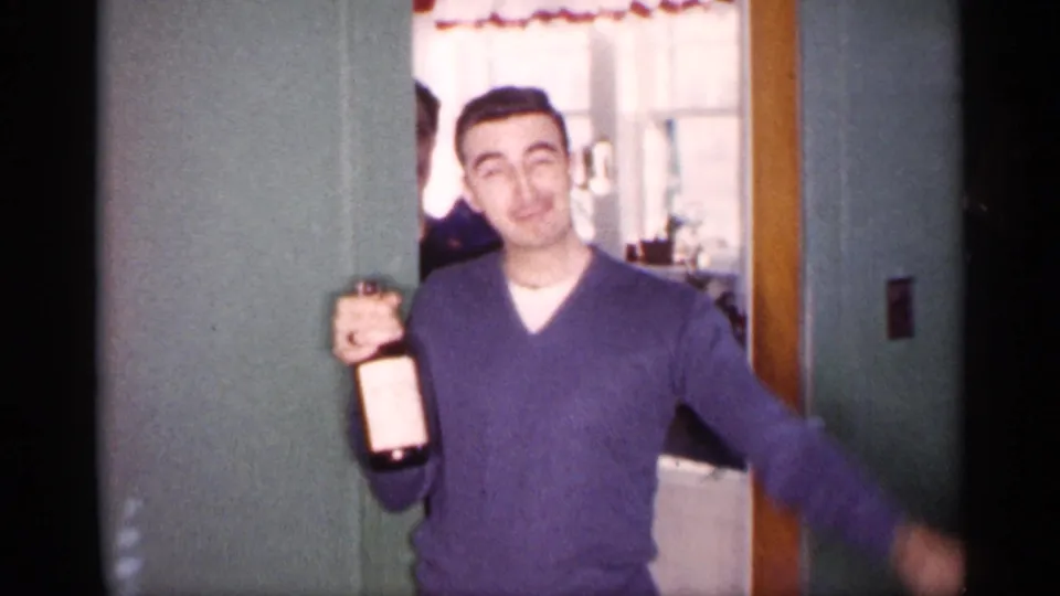 The young man stands near the doorway holding a bottle toward the camera, with green walls and a curtained kitchen or adjoining room behind him.