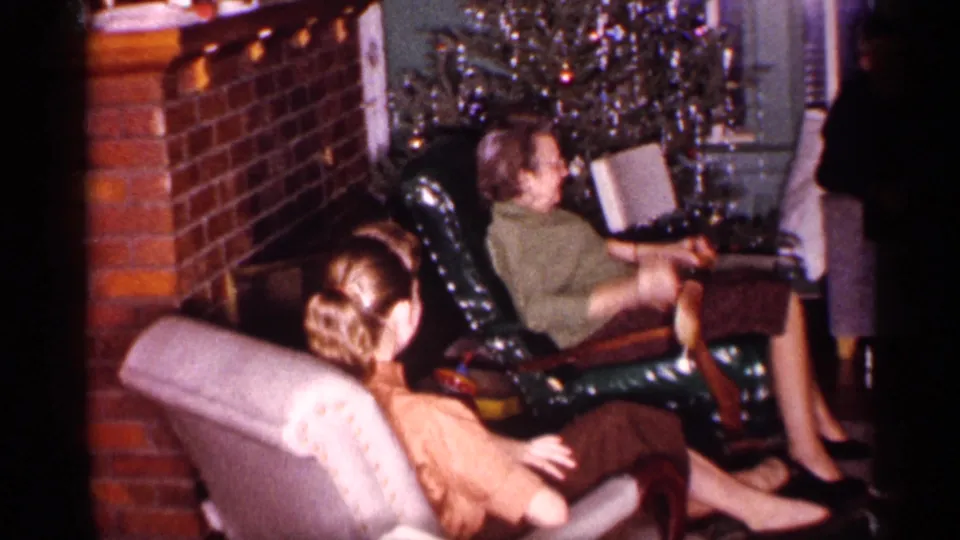 Adults sit in upholstered chairs near a brick fireplace and decorated Christmas tree during a 1959 Michigan home-movie holiday gathering.