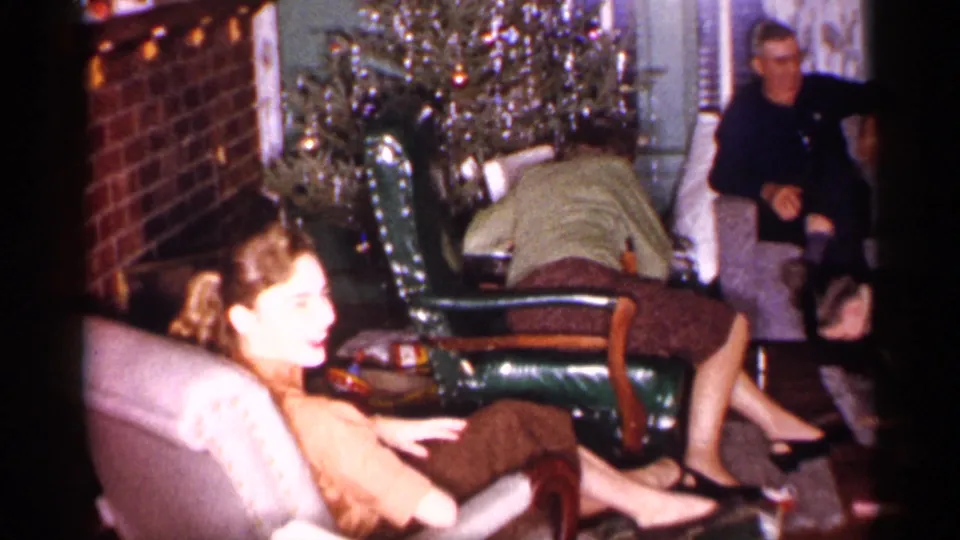 A woman faces the camera while other adults sit or bend near chairs by a Christmas tree in a private Michigan living room.
