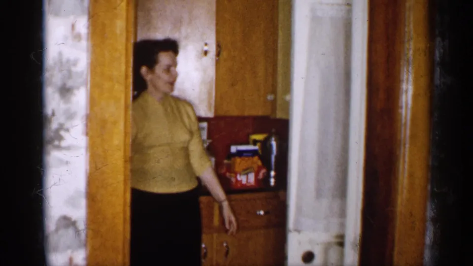 1959 Michigan home-movie frame of a woman in a yellow sweater reaching near wood kitchen cabinets with visible pantry packaging.
