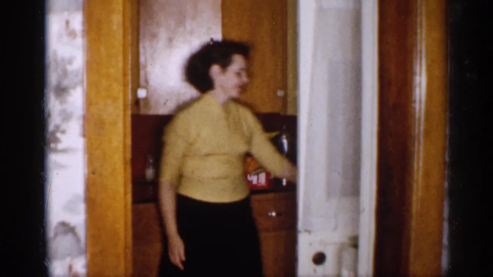 1959 Michigan home-movie frame showing a woman in a yellow sweater moving through a warm wood kitchen interior.