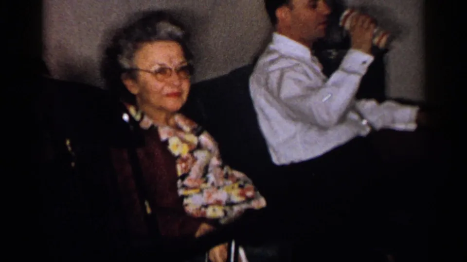 1959 Michigan home-movie frame showing an older woman with glasses seated beside a man drinking from a can in an indoor setting.
