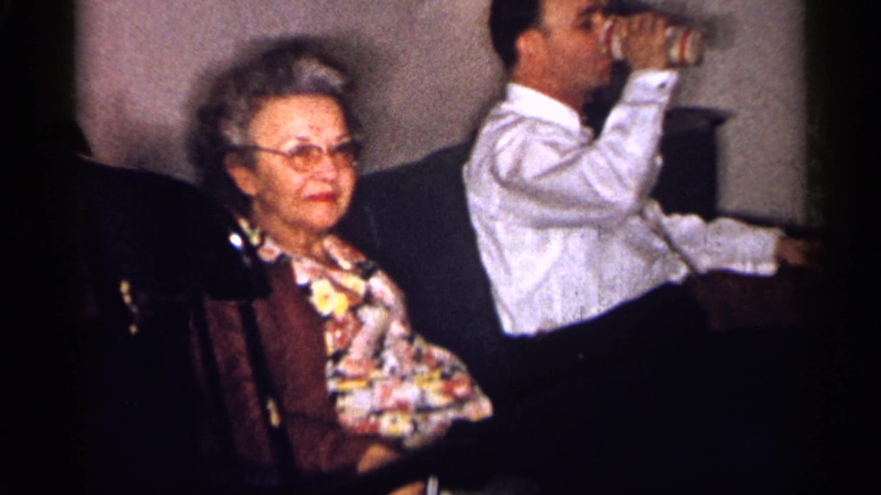 Older woman with glasses sits on a dark couch beside a man drinking from a glass.