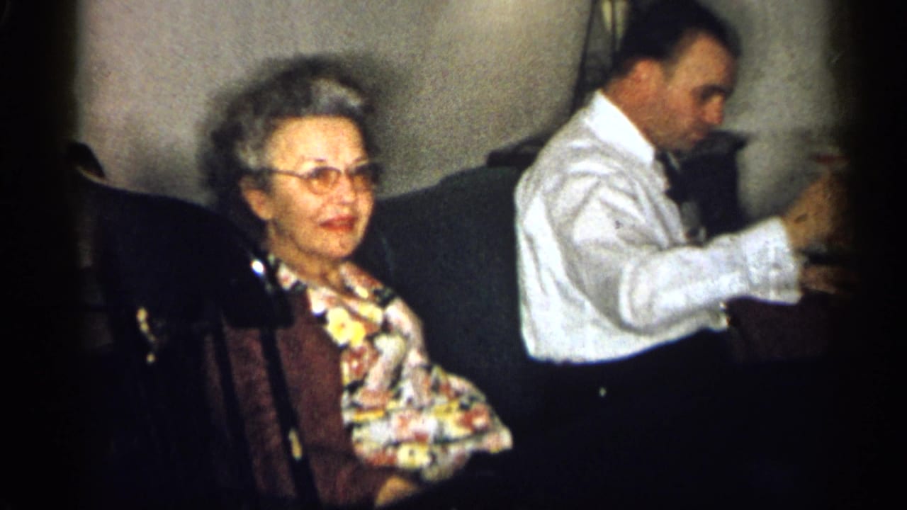 1959 Michigan interior frame with an older woman on a couch and a man in a white shirt holding a glass beside her.