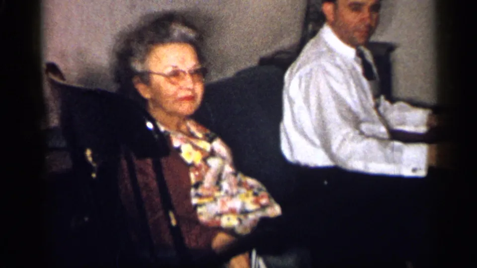 1959 Michigan home-movie still of an older woman in glasses seated beside a man in a white shirt and tie looking toward camera.