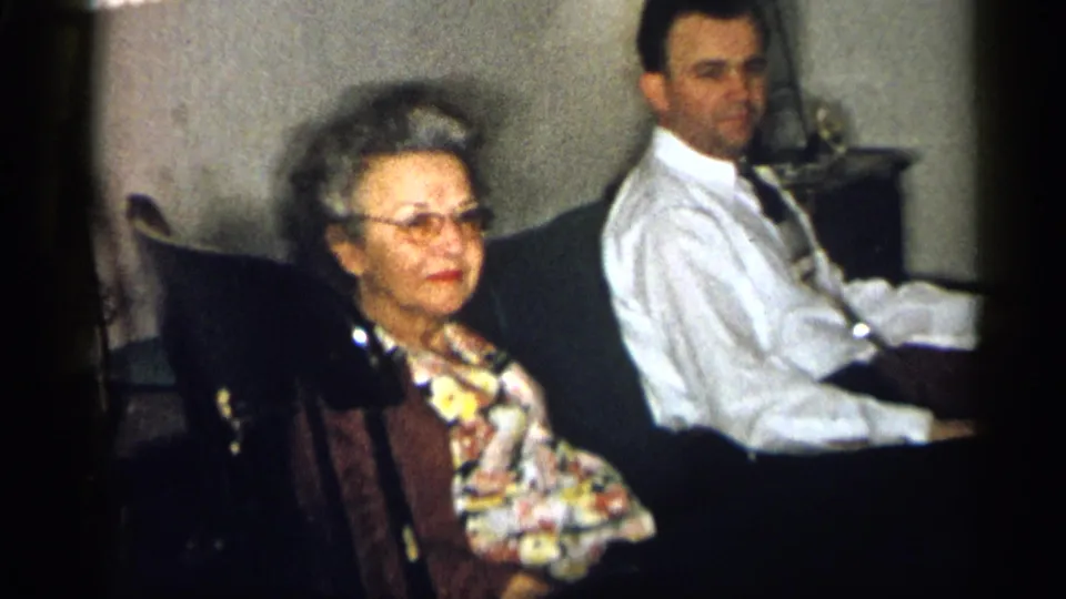 1959 Michigan frame showing an older woman with glasses and a man in a white shirt seated together in a private living room.