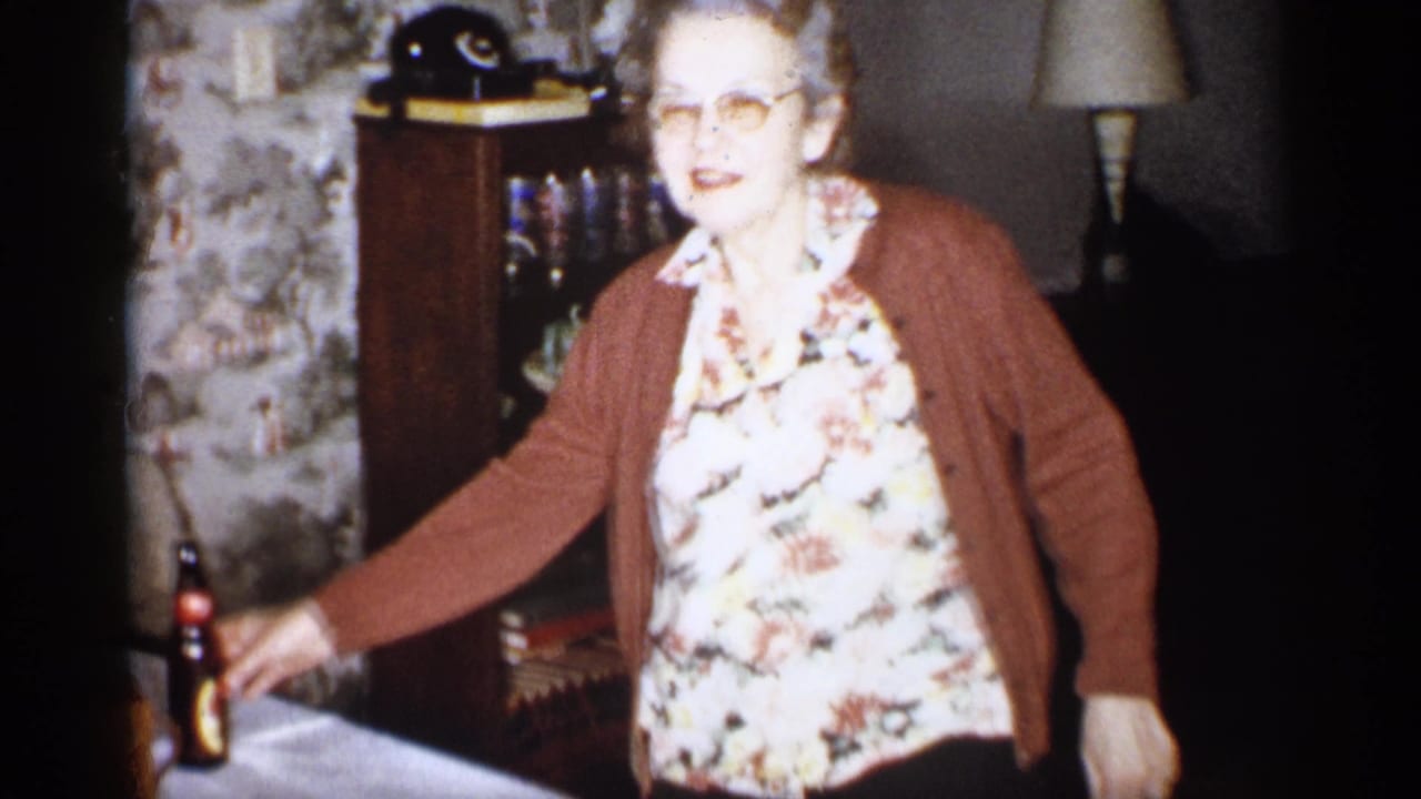Older woman in glasses stands beside a table holding a dark bottle in a vintage living room.
