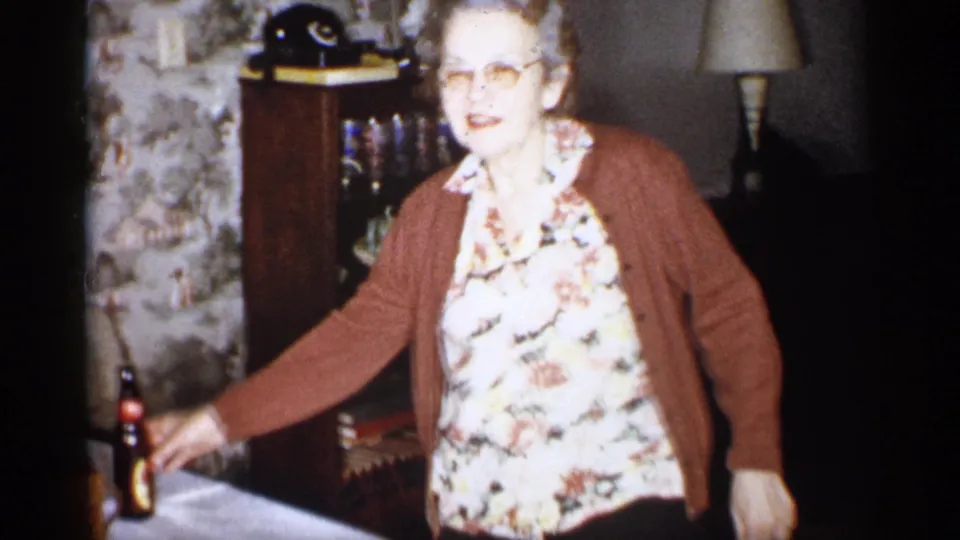 In a 1959 Michigan home-movie frame, an older woman in glasses, a floral blouse, and a brown cardigan stands in a patterned-wallpaper living room beside a table with a dark glass bottle.