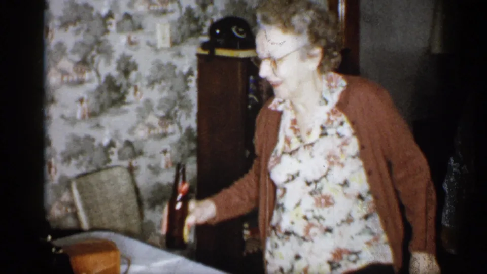 The older woman leans toward a table and reaches for a dark glass bottle in a Michigan living room with patterned wallpaper, a chair, cabinet, and black rotary telephone visible behind her.