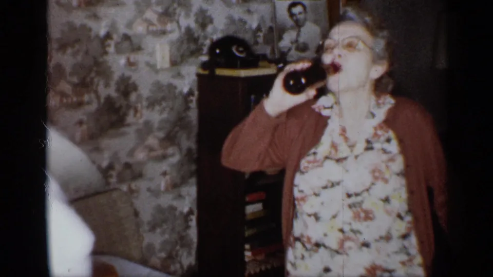 The woman lifts the dark glass bottle to her mouth while standing in the living room, with another adult passing close to the camera and partially obscuring the left side of the frame.