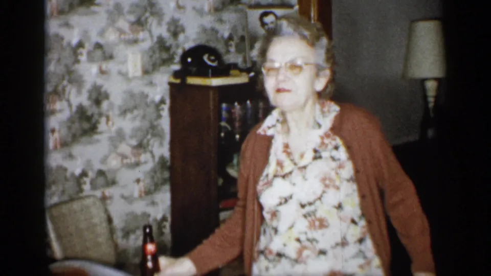 The older woman stands with the dark glass bottle lowered at her side, facing the camera in the 1959 Michigan living room with patterned wallpaper, cabinet, rotary telephone, chair, and lamp nearby.