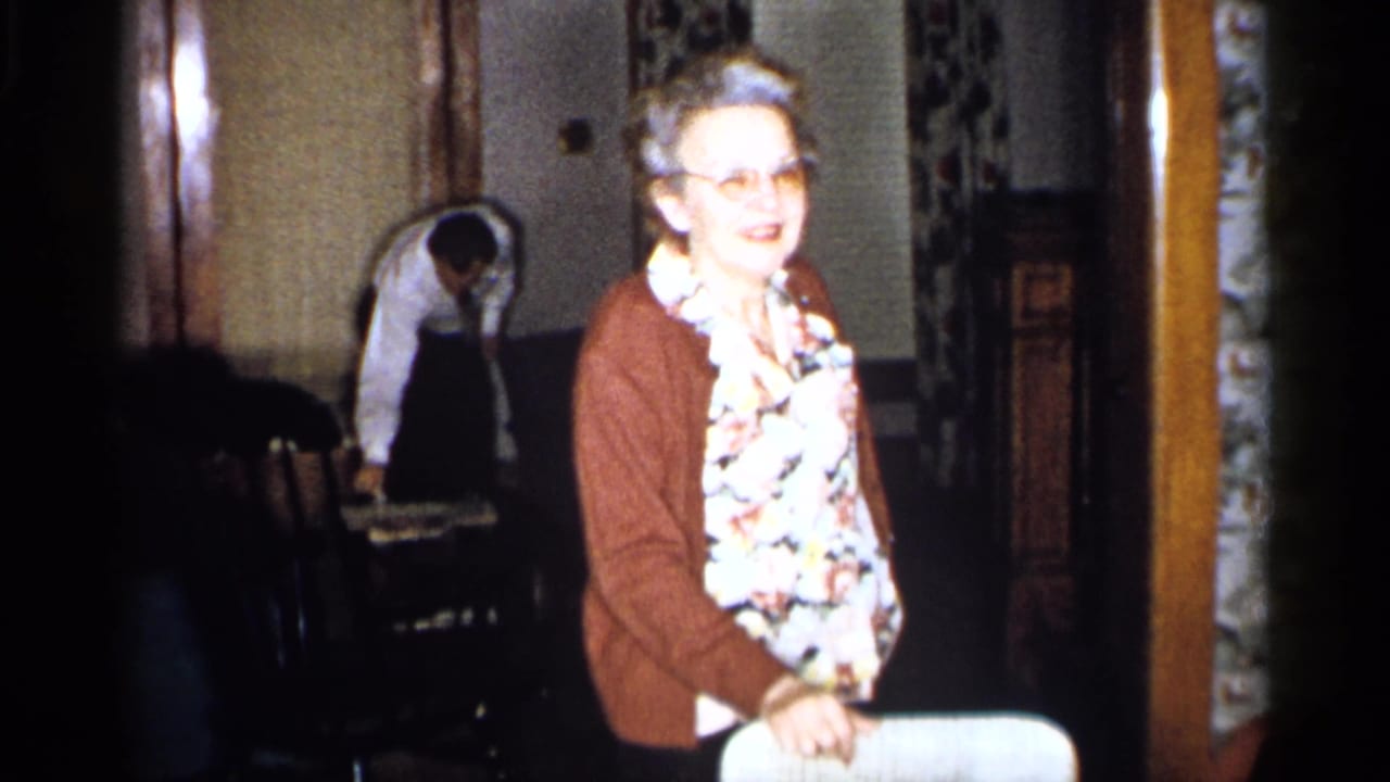 Older woman in a brown cardigan smiles beside a white chair in a home interior.