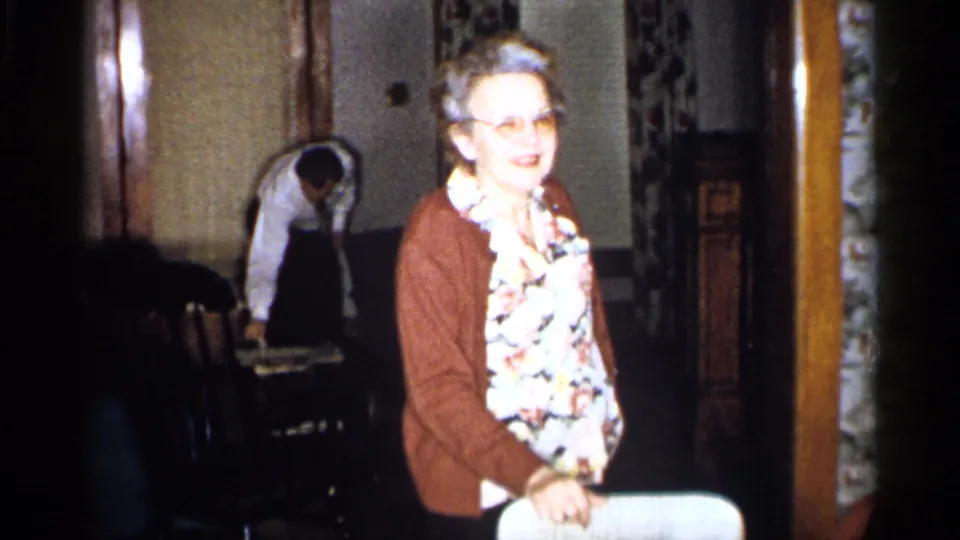 In a 1959 Michigan home-movie frame, an older woman in a floral blouse and brown cardigan stands near a white chair as another woman bends in the background.