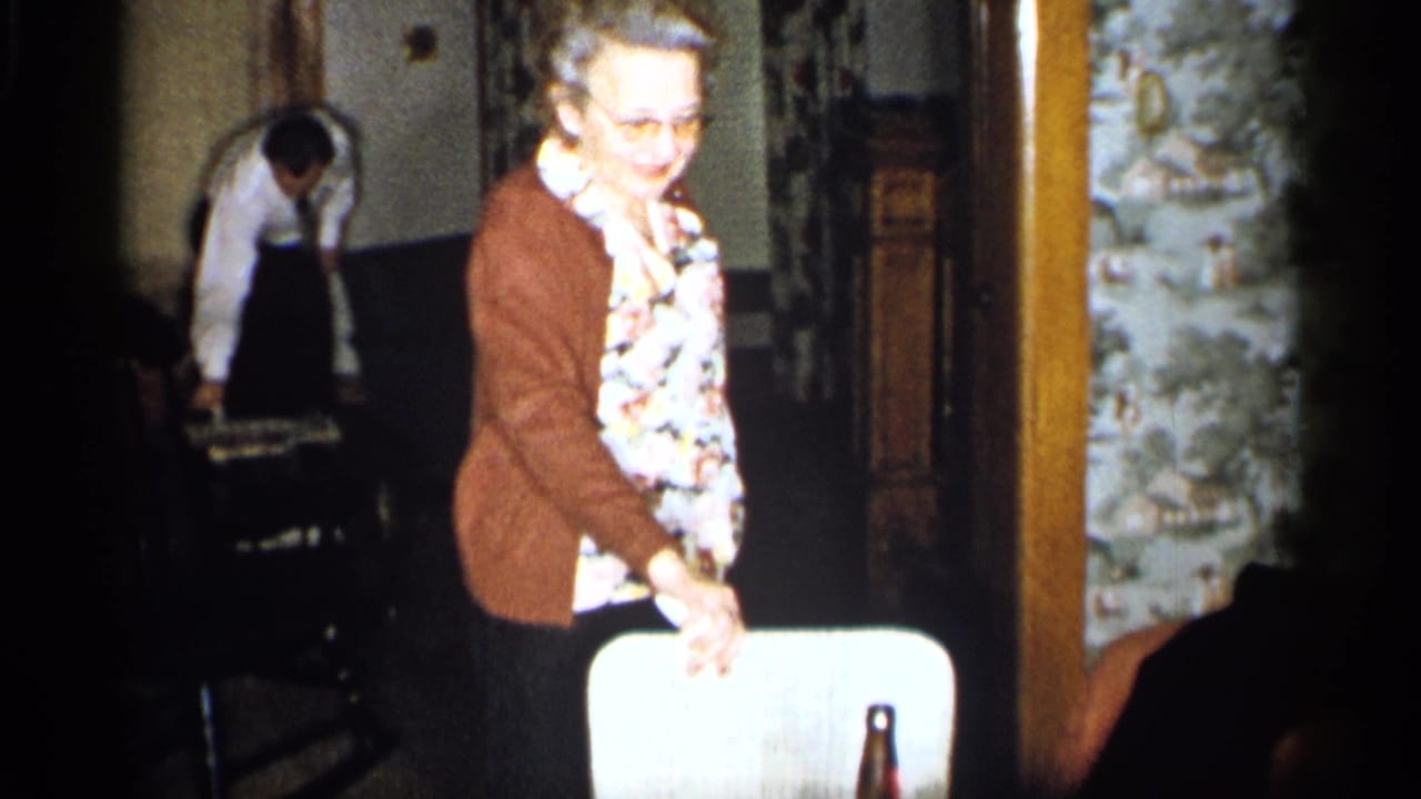 Older woman in cardigan stands by a white chair with a bottle visible on a table.