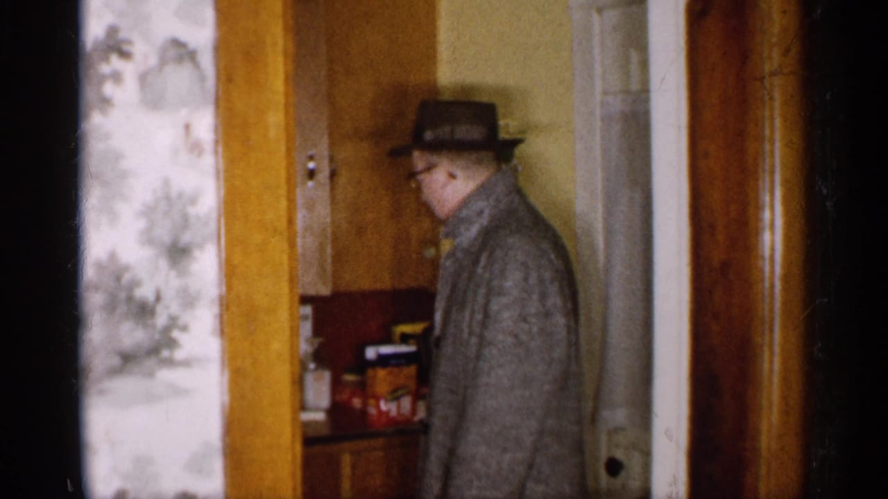 Man in a fedora and gray coat standing in a home doorway.