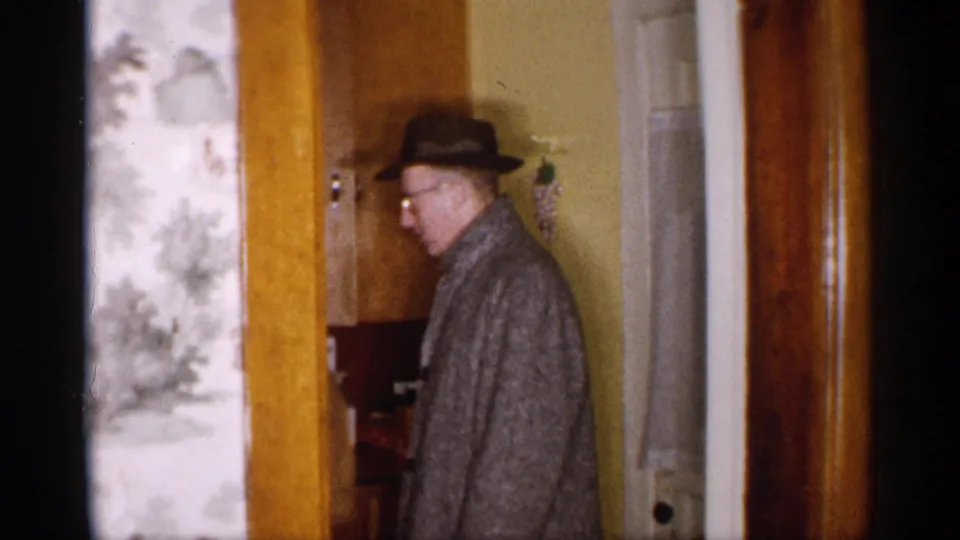 A man in a fedora and gray coat turns near a doorway inside a 1959 Michigan private home.