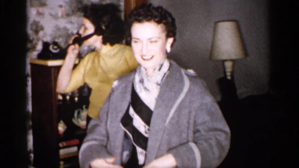 A smiling woman in a gray coat and patterned scarf stands inside a 1959 Michigan home as another woman uses a rotary telephone.