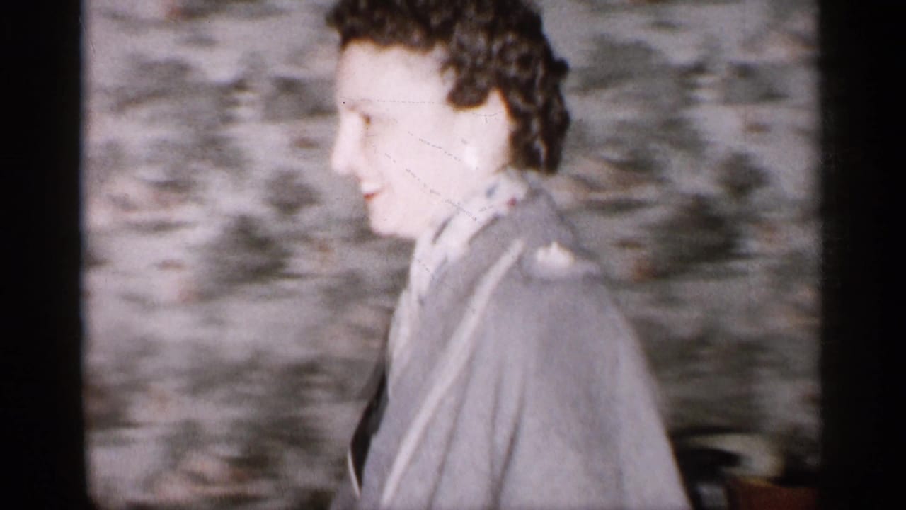 In a 1959 Michigan home-movie frame, a woman in a gray coat and scarf smiles beside patterned wallpaper near a doorway.