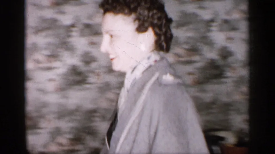 In a 1959 Michigan home-movie frame, a woman in a gray coat and scarf smiles beside patterned wallpaper near a doorway.