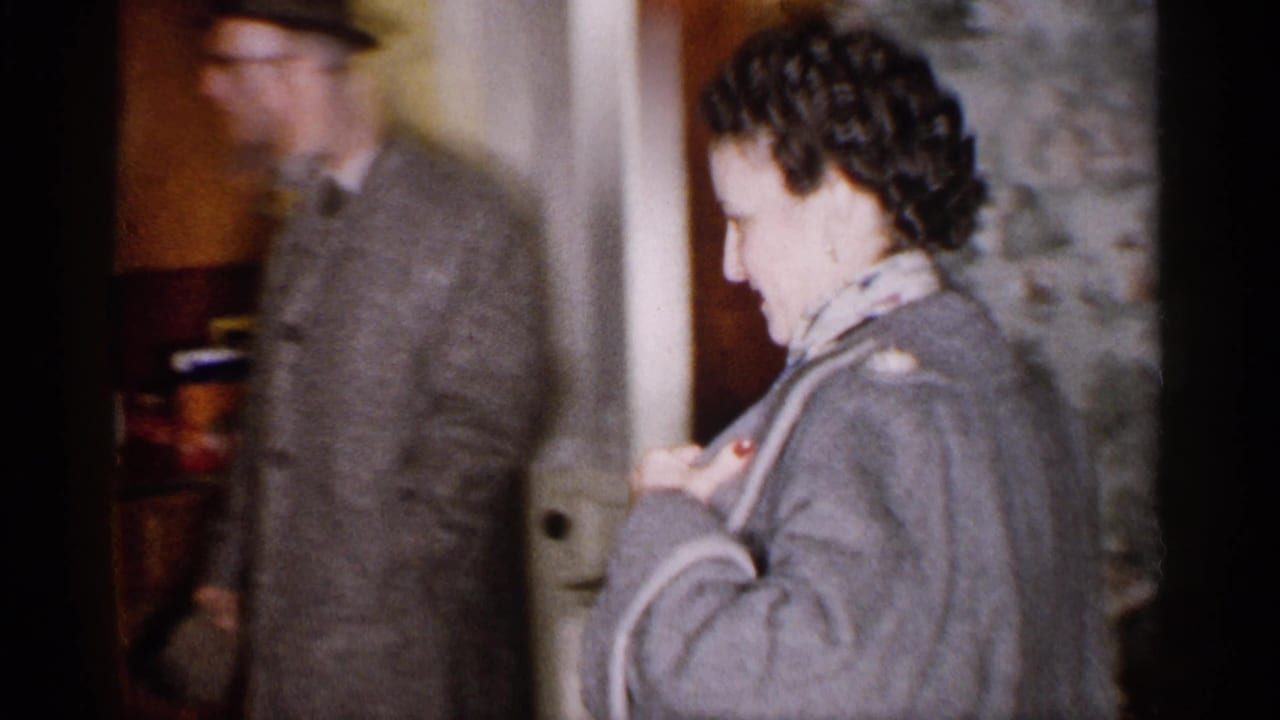 Woman in a gray coat stands near a doorway as a man in a hat follows behind.