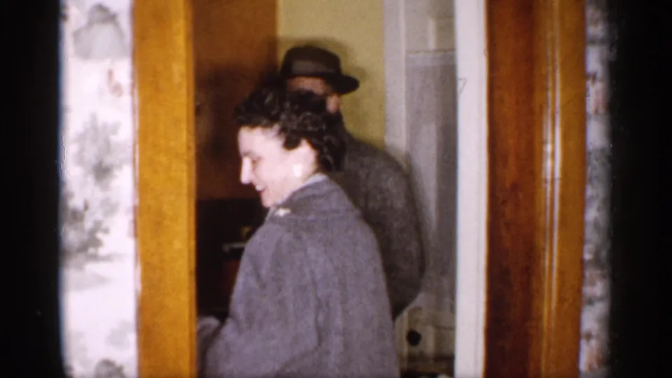 The woman turns with a smile near the wood-trimmed doorway while the man remains partly visible behind her in the private home interior.