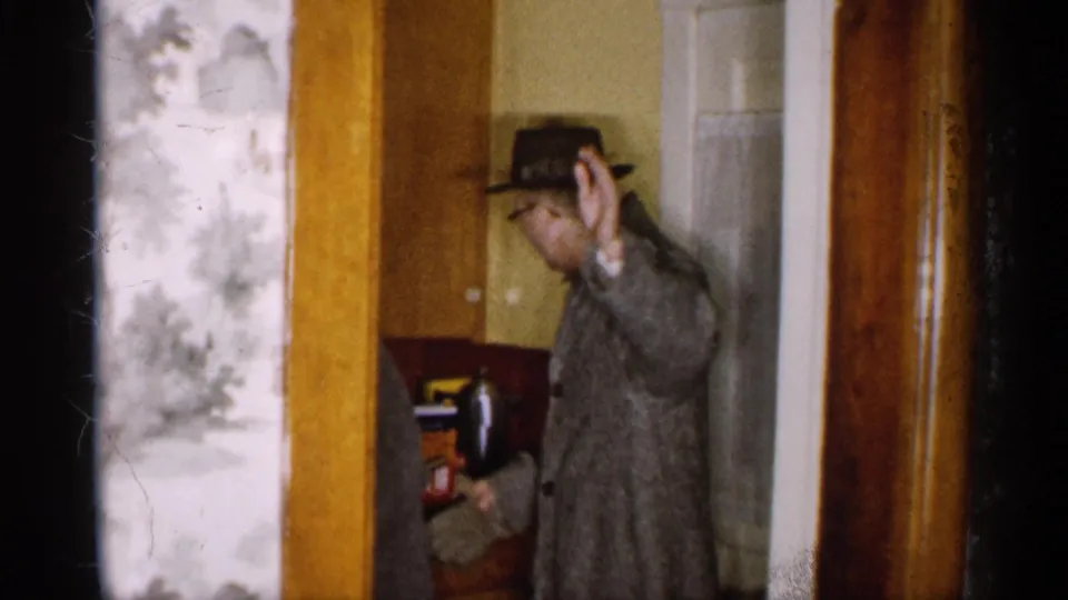 A man in a brimmed hat and overcoat waves from the doorway of a Michigan home, with wood trim and interior furnishings visible.