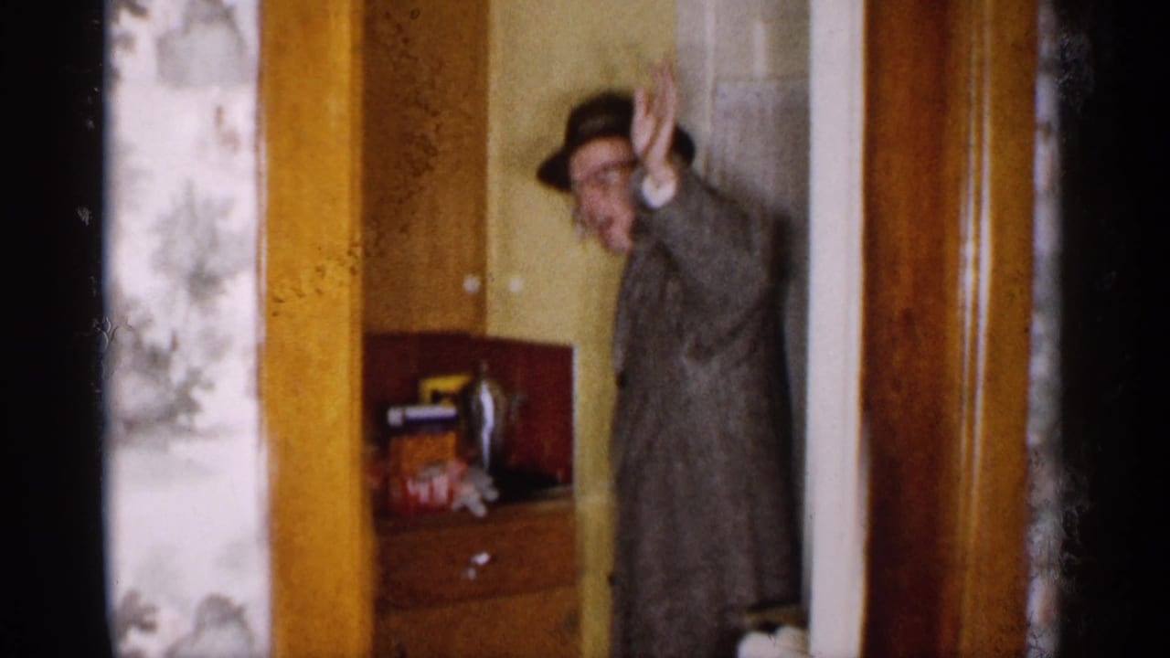 Man in a fedora and coat raises his hand from a kitchen doorway.