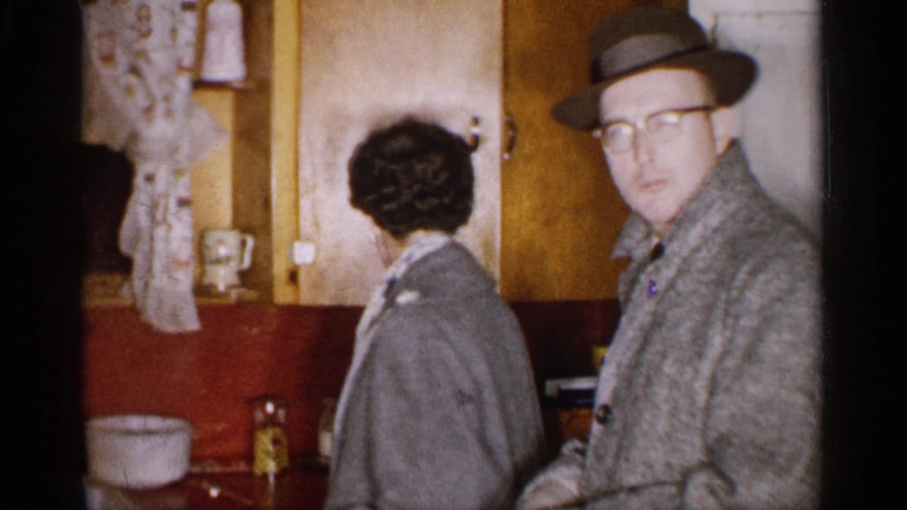 A woman in a coat stands beside a man in a fedora and glasses in a warm-toned Michigan kitchen interior.