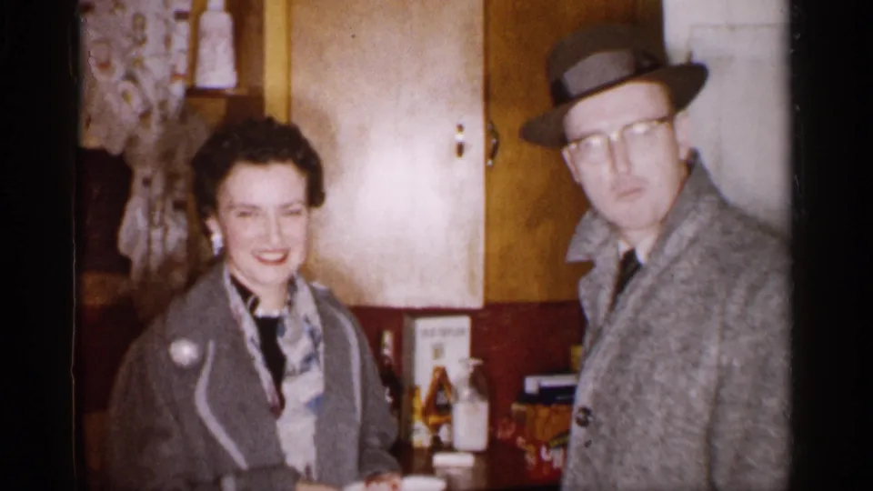 A smiling woman and a man in a fedora pose in a Michigan kitchen, captured in warm 1959 8mm home-movie color.