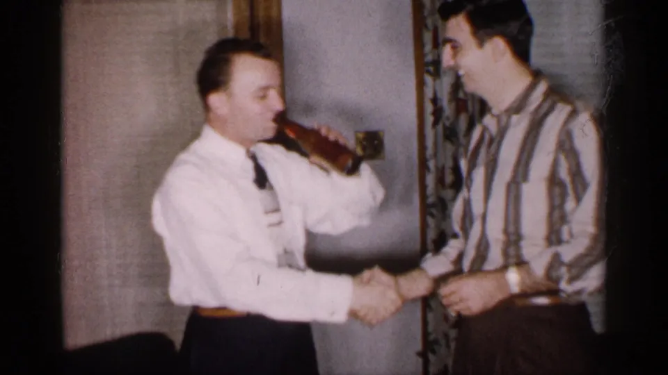 The man in the white shirt and tie drinks from the bottle while shaking hands with a smiling man in a striped shirt.