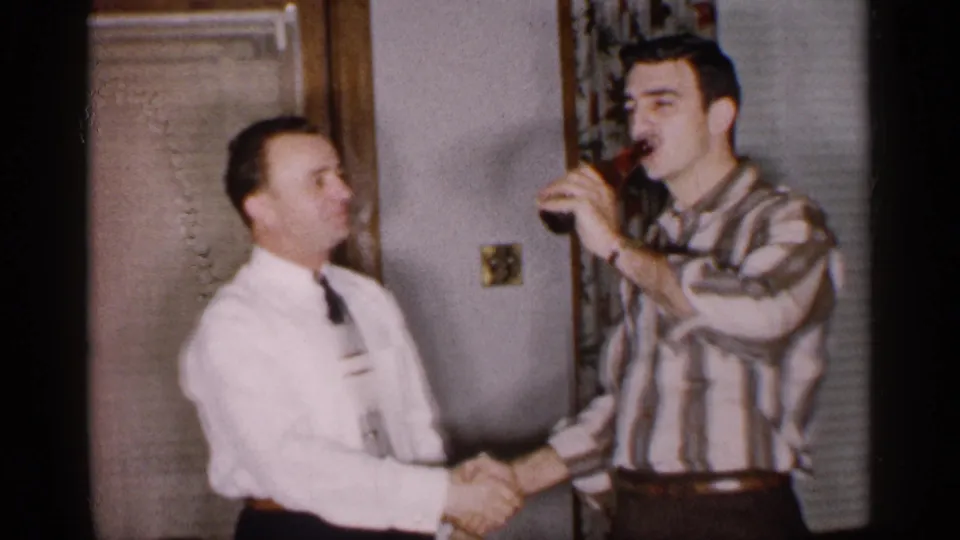 The bottle has passed to the man in the striped shirt, who drinks while the man in the white shirt maintains the handshake.