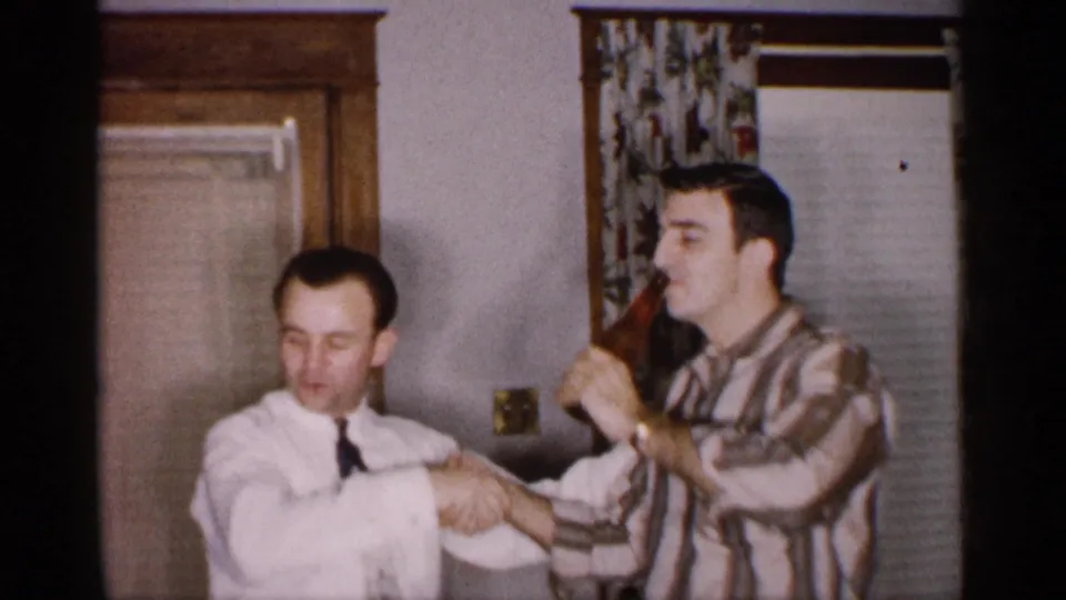 Inside a Michigan home setting, the man in the striped shirt drinks from the brown bottle while both men keep shaking hands.