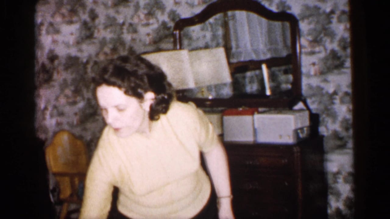Woman in a yellow sweater moves through a room with patterned wallpaper and a dresser mirror.