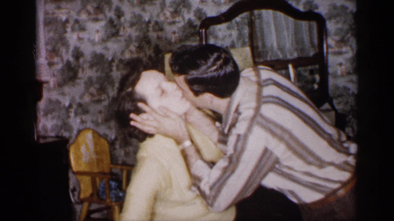 A man in a striped shirt embraces and kisses a woman in a yellow sweater inside the 1959 Michigan home setting.