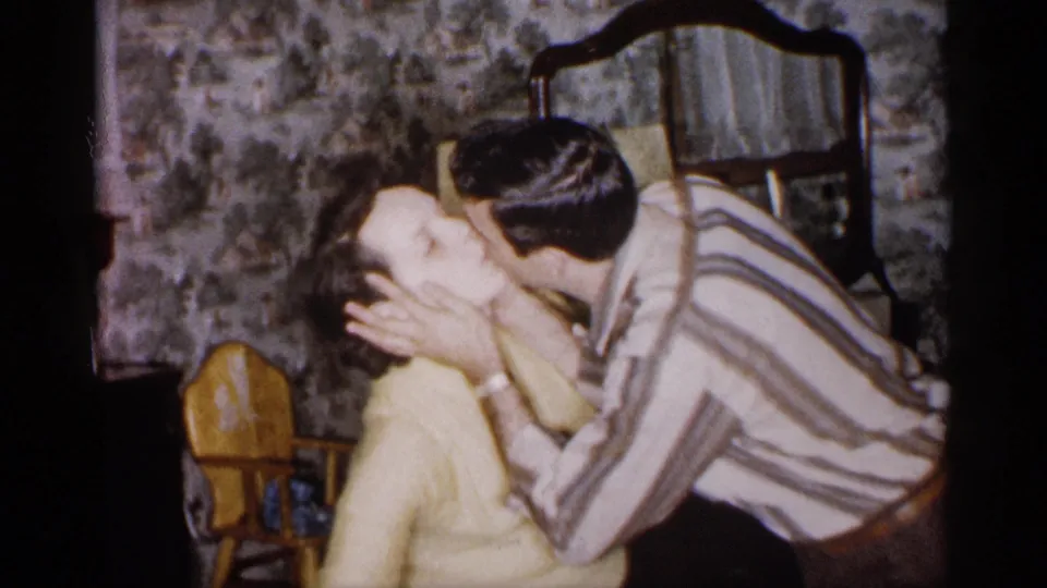 A man in a striped shirt embraces and kisses a woman in a yellow sweater inside the 1959 Michigan home setting.