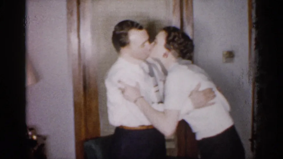 Two adult men in dress shirts and ties share a close affectionate greeting beside a wooden door in the 1959 Michigan home movie.
