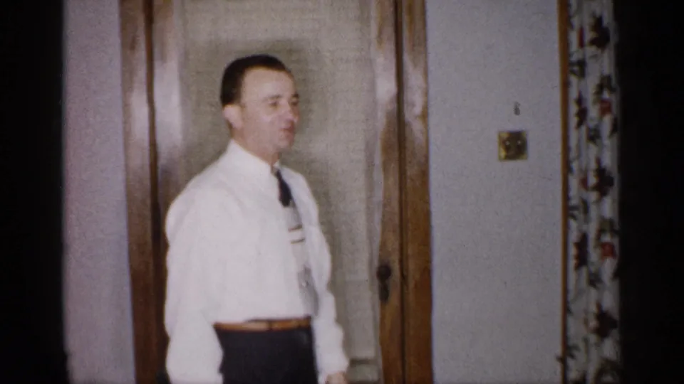 A man in a white shirt and tie stands in the Michigan home interior and appears to wave or gesture toward the camera.