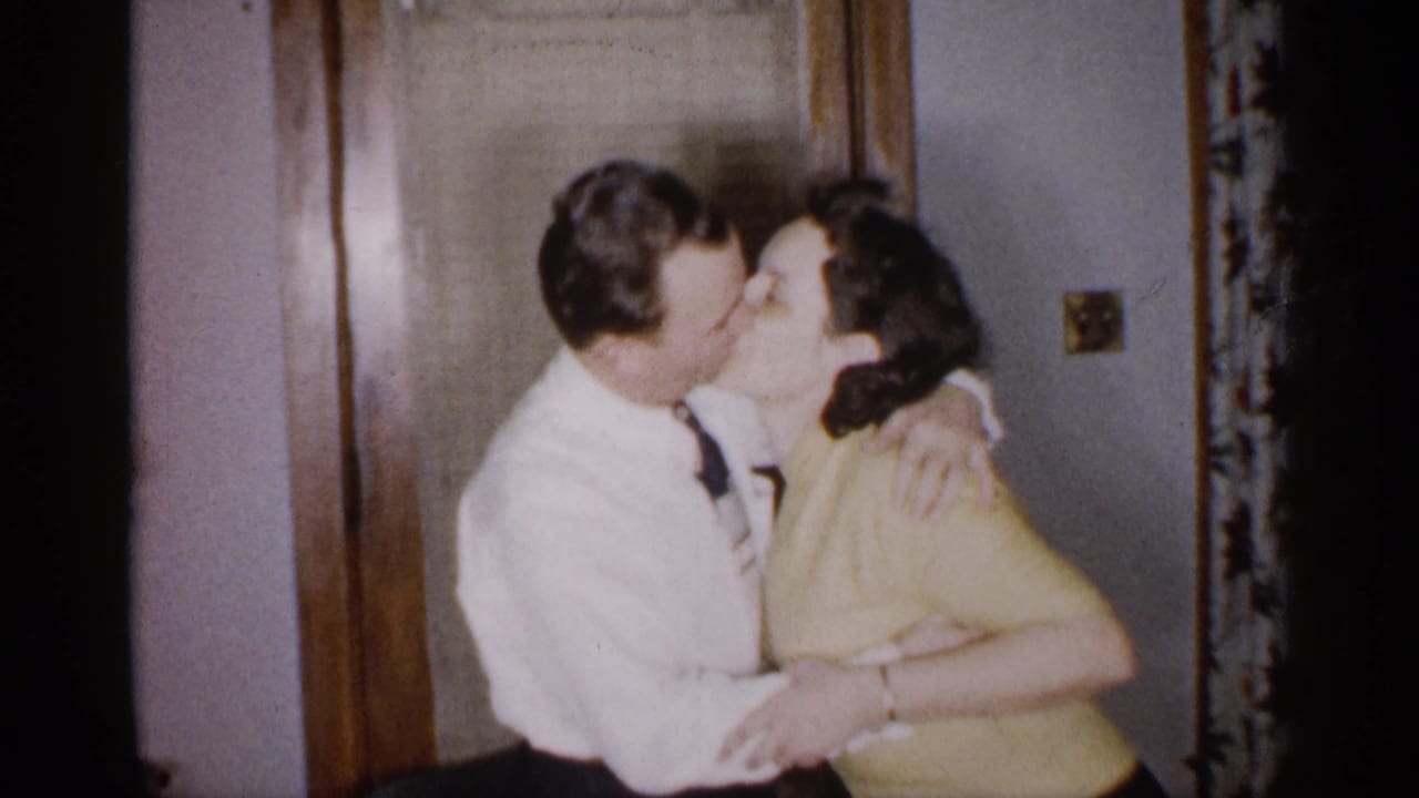 In a 1959 Michigan home-movie frame, an adult man in a white dress shirt and tie embraces and kisses an adult woman in a yellow top near a wooden interior door.