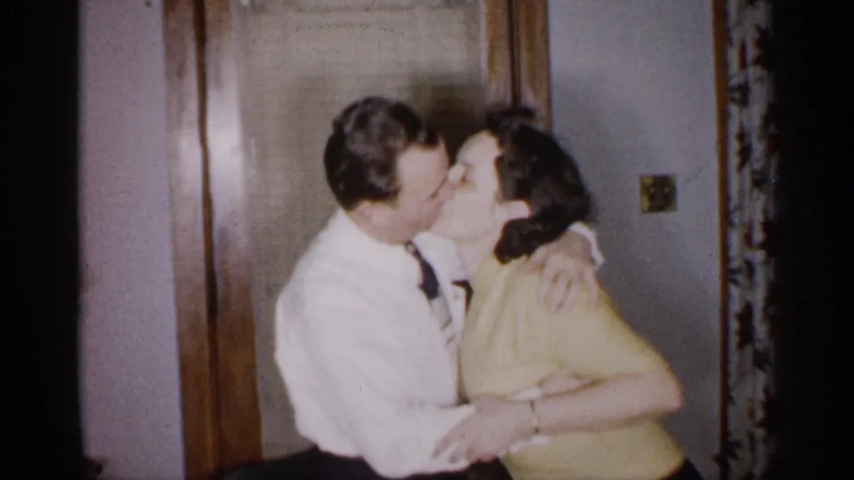 In a 1959 Michigan home-movie frame, an adult man in a white dress shirt and tie embraces and kisses an adult woman in a yellow top near a wooden interior door.